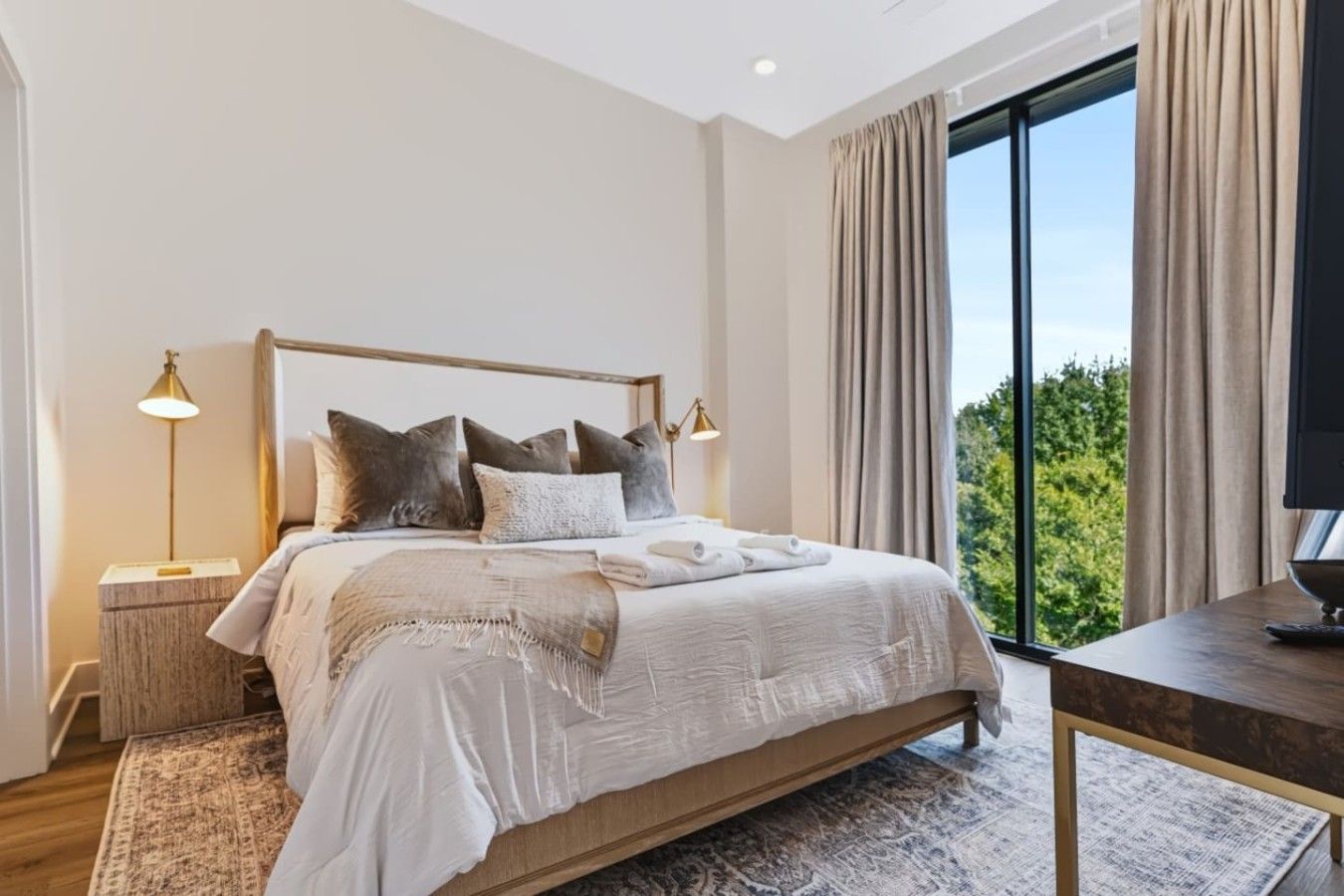 Une chambre moderne avec un grand lit, des lampes de chevet, une vue sur la verdure par la fenêtre et une petite table.
