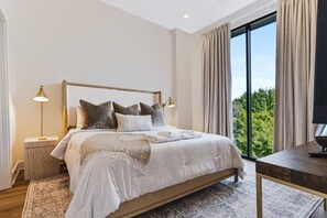 Un dormitorio moderno con una cama grande, lámparas de noche, vista a zonas verdes por la ventana y una mesita.