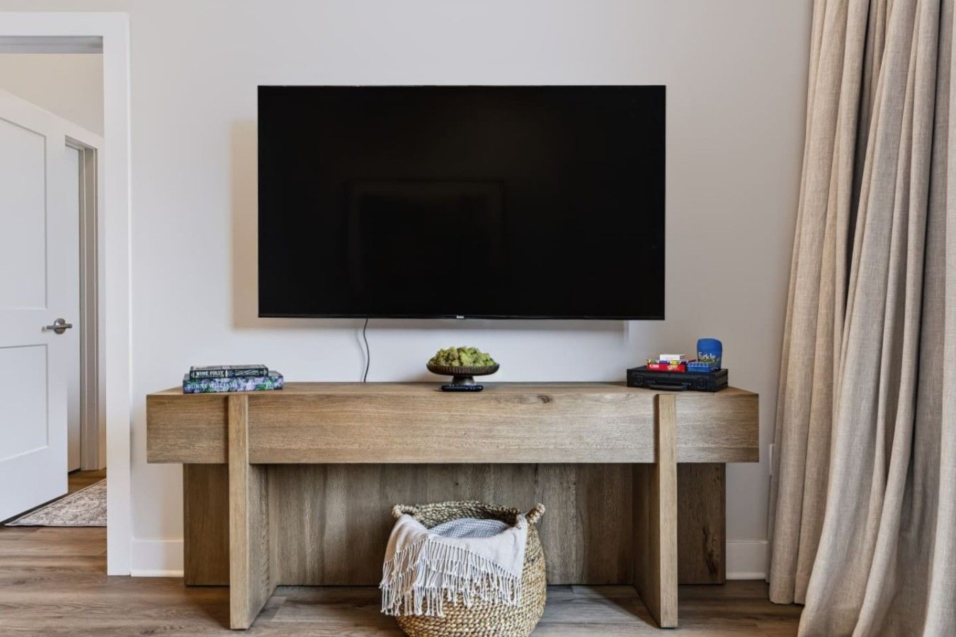 A room with a flat-screen TV mounted on a wooden console table, a woven basket with a blanket, and a small shelf with books and toys.