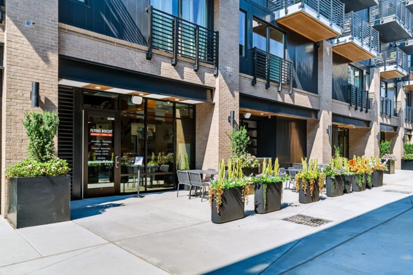 Un bâtiment moderne abritant un restaurant au rez-de-chaussée, doté d’une terrasse et agrémenté de jardinières verdoyantes.