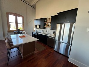 A modern kitchen with a central island, dark cabinetry, stainless steel appliances, and a view of the city through a large window. - City Views & Comfort | Heart of Downtown Knoxville (Knoxville)