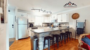 A modern kitchen with white cabinets, a central island with bar stools, a stone accent wall, and a clock on the wall. - Powdered Sugar at Shallowford Walk (Sugar Mountain)