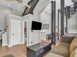 A modern living room with a flat-screen TV, a coffee table, and a sofa. There is a kitchen area with a dining table and chairs visible through an open doorway. - The Jackson Gem | Steps to Knoxville’s Old City (Knoxville)