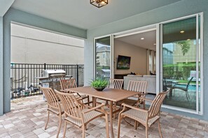 An outdoor patio with a wooden table and chairs, a grill, and a view of a swimming pool and a cityscape through a glass door. - 6BR Near Disney Private Pool, spa & Loft (Kissimmee)
