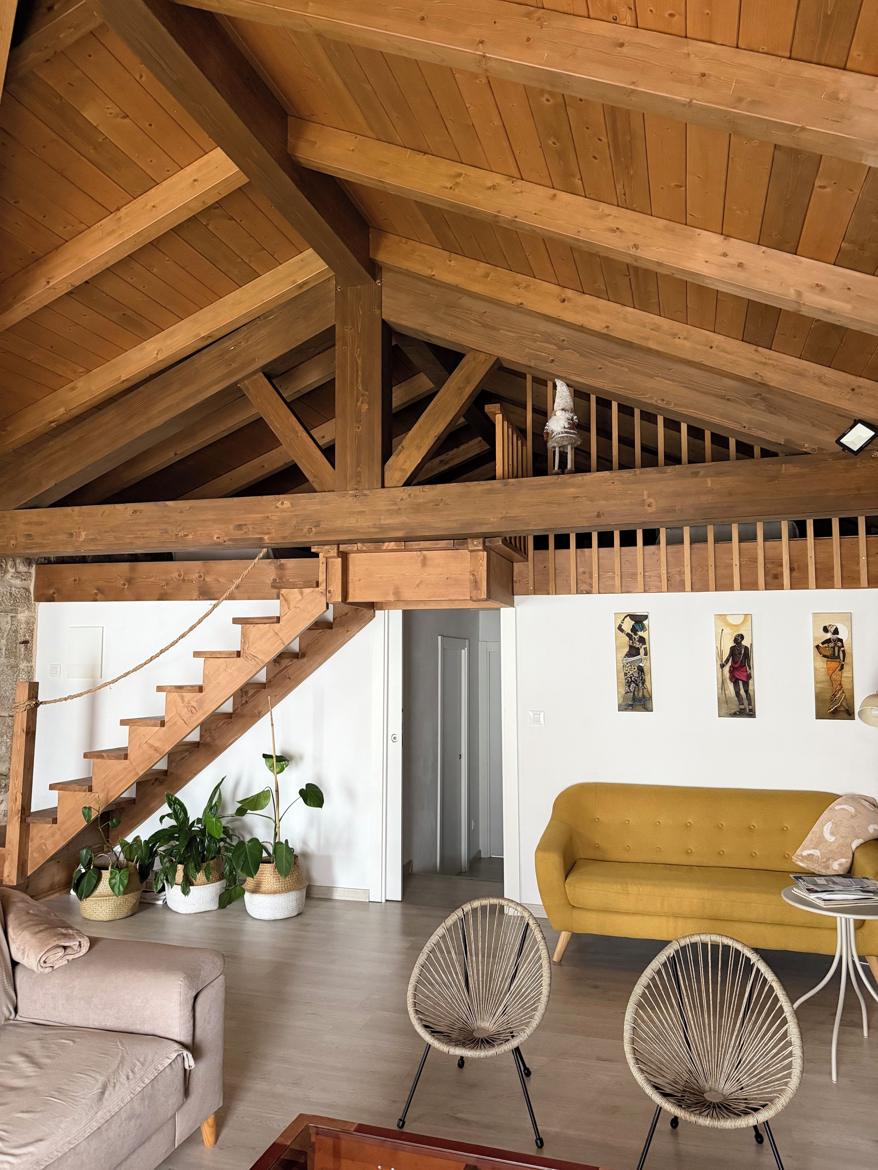 Un loft amplio con techo alto, vigas de madera a la vista, una escalera, un sofá, dos sillones de mimbre y obras de arte en la pared.