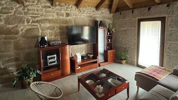 Een rustieke woonkamer met stenen muren, houten balken, een stenen open haard, een televisie en een bank.