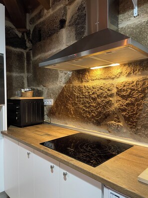 A kitchen with a stone wall backsplash, a stainless steel range hood, a black induction cooktop, and a microwave. - As Fontaiñas (Berán)