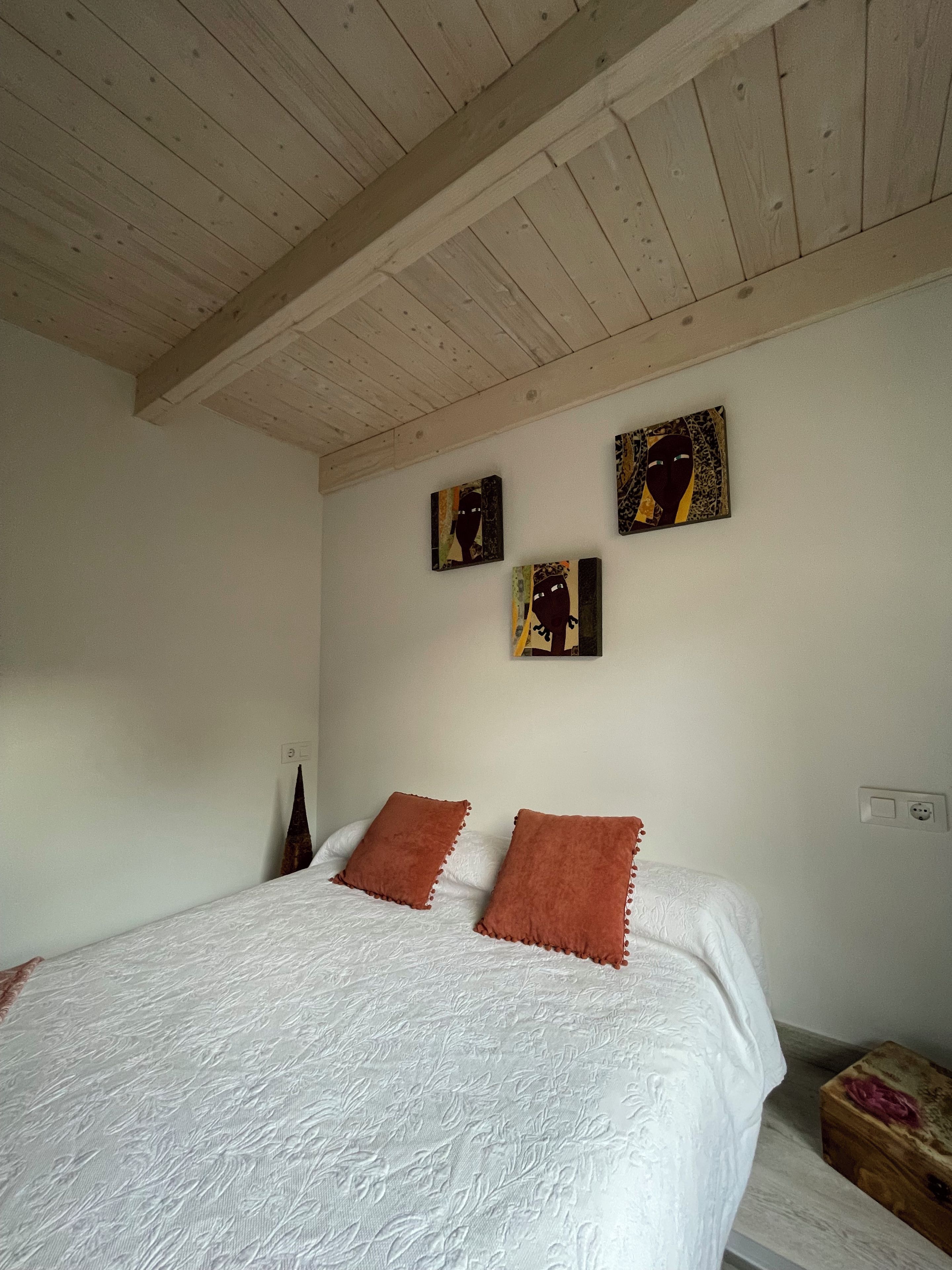 Une chambre avec un plafond en bois, un lit avec une literie blanche et deux oreillers marron, et trois œuvres d’art accrochées au mur.
