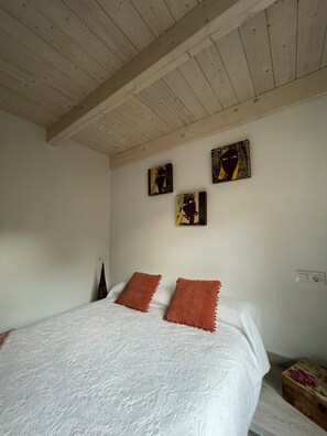 Une chambre avec un plafond en bois, un lit avec une literie blanche et deux oreillers marron, et trois œuvres d’art accrochées au mur.