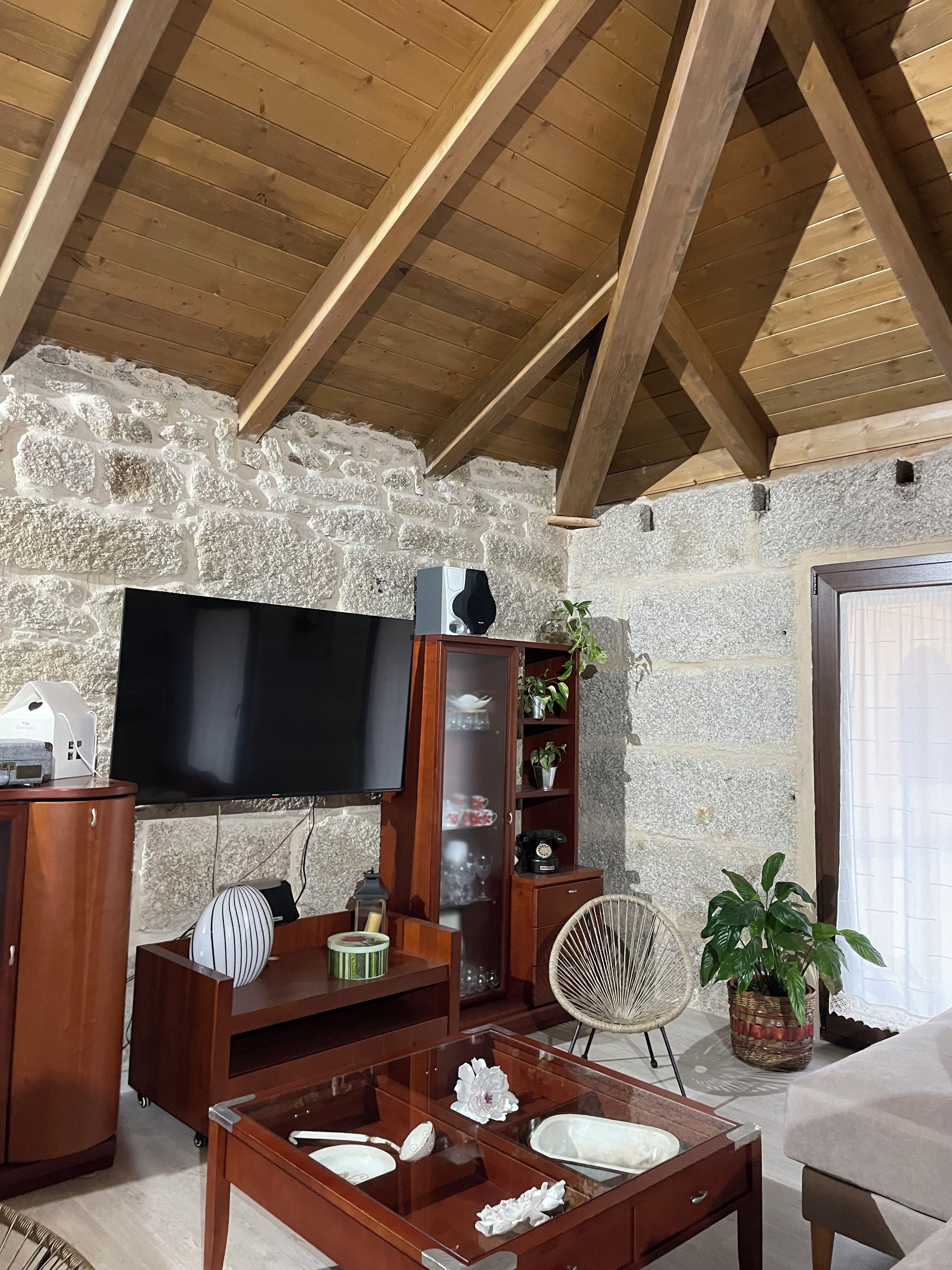 Un salon chaleureux avec un mur en pierre, des poutres apparentes en bois au plafond, un téléviseur à écran plat, un meuble en bois, une table basse en verre agrémentée d’objets décoratifs et un fauteuil en osier.