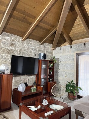Un salon chaleureux avec un mur en pierre, des poutres apparentes en bois au plafond, un téléviseur à écran plat, un meuble en bois, une table basse en verre agrémentée d’objets décoratifs et un fauteuil en osier.