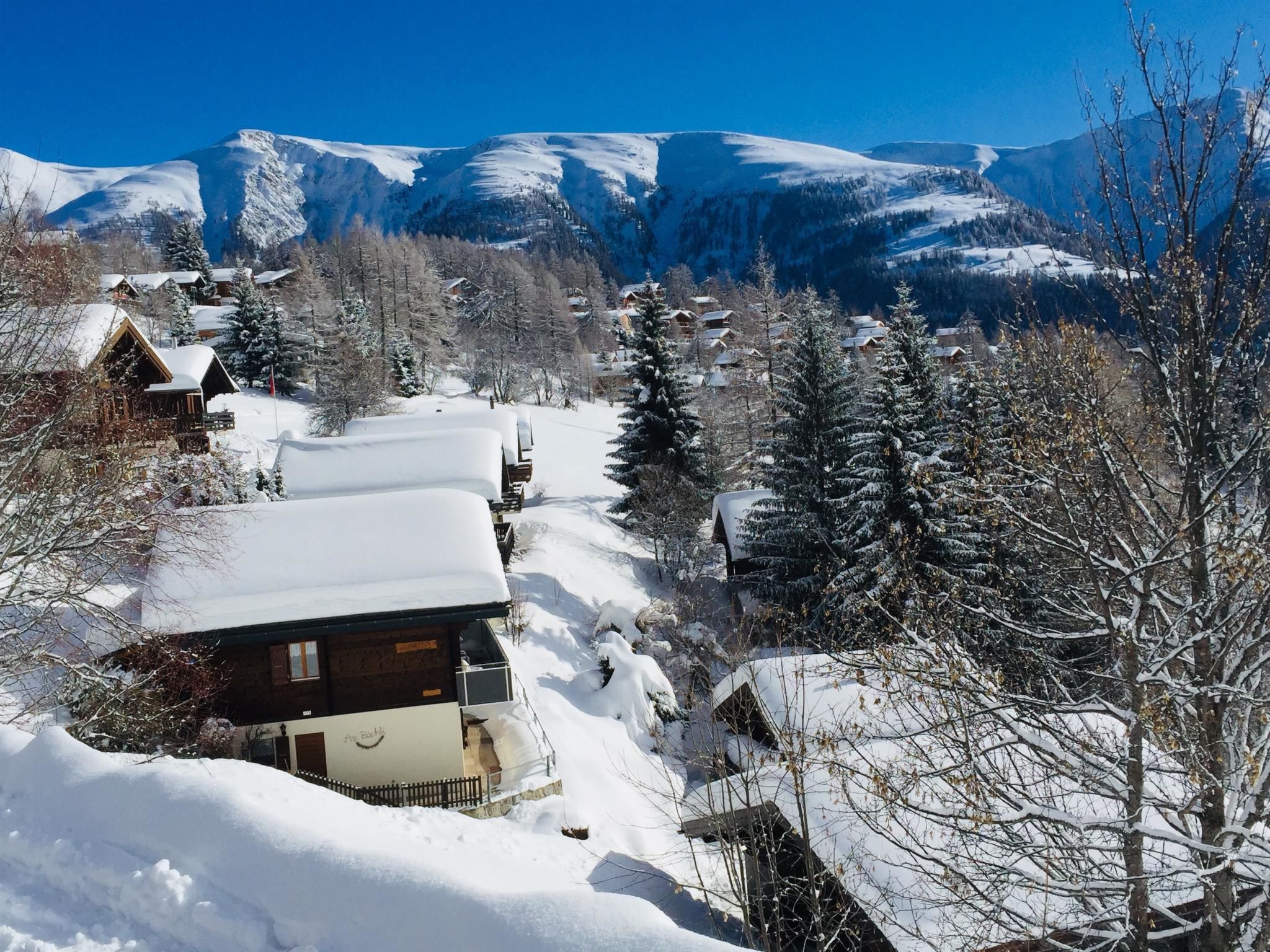 A snow-covered village with wooden houses, a clear sky, and mountains in the background.