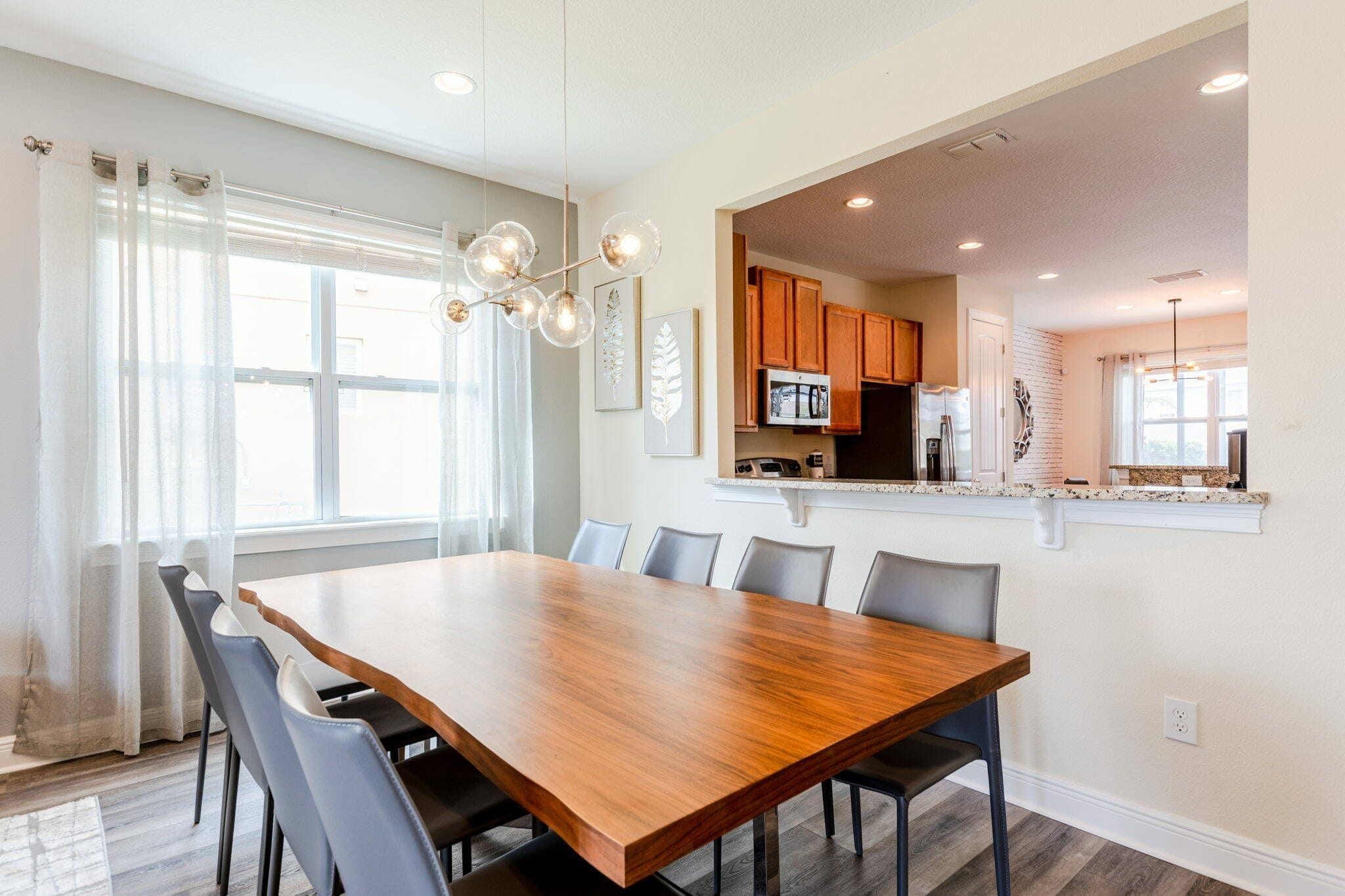 A dining area with a wooden table and chairs, a kitchen with wooden cabinets, and a window with sheer curtains.