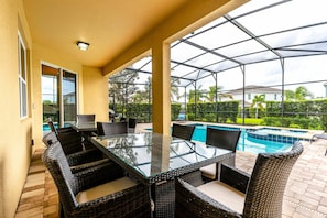 A covered outdoor patio with a glass-top table and wicker chairs, overlooking a swimming pool. - 6BR Near Disney Private Pool, spa & Game Room (Kissimmee)
