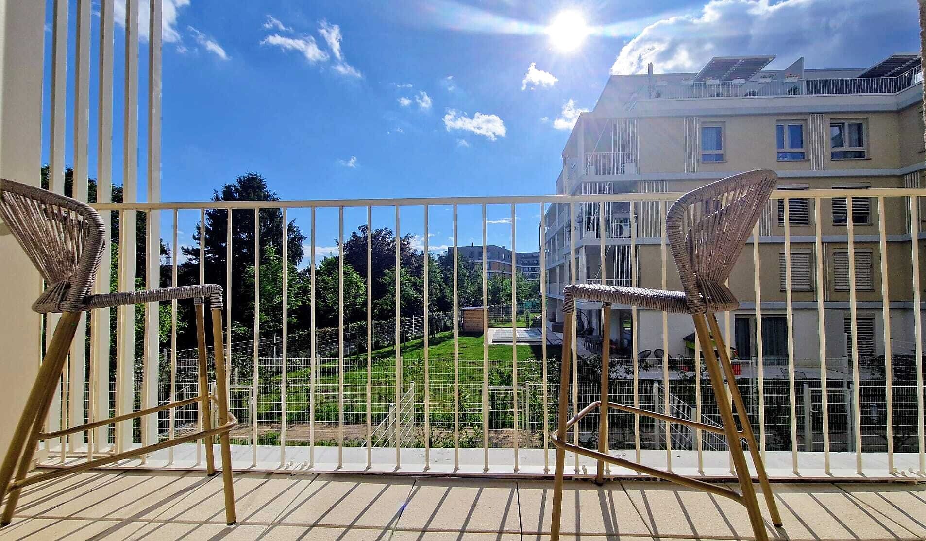 A balcony with two wicker chairs, a metal railing, and a view of a green park and buildings.