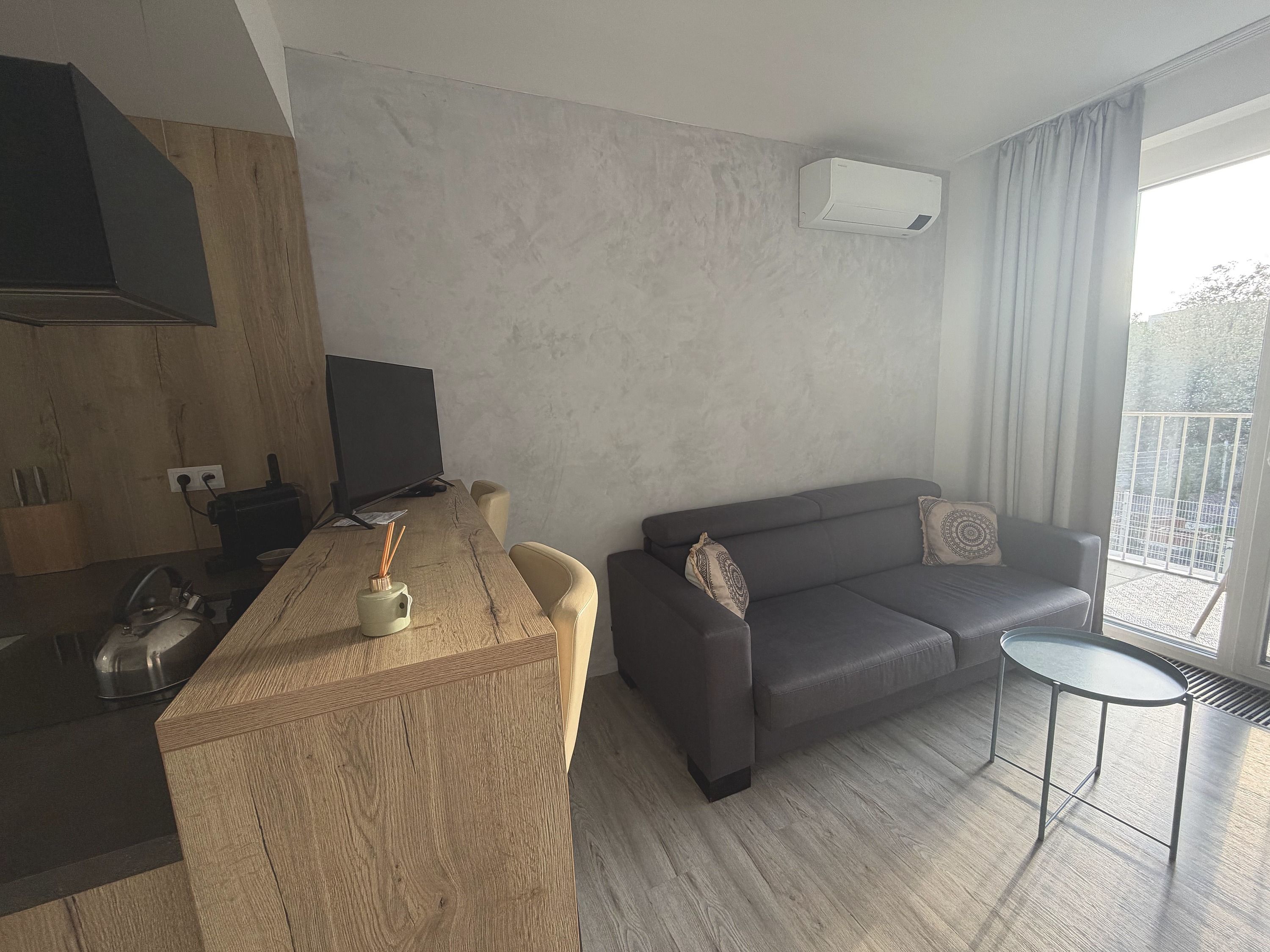 En moderne stue med et køkkenområde, en grå sofa og et lille rundt bord.