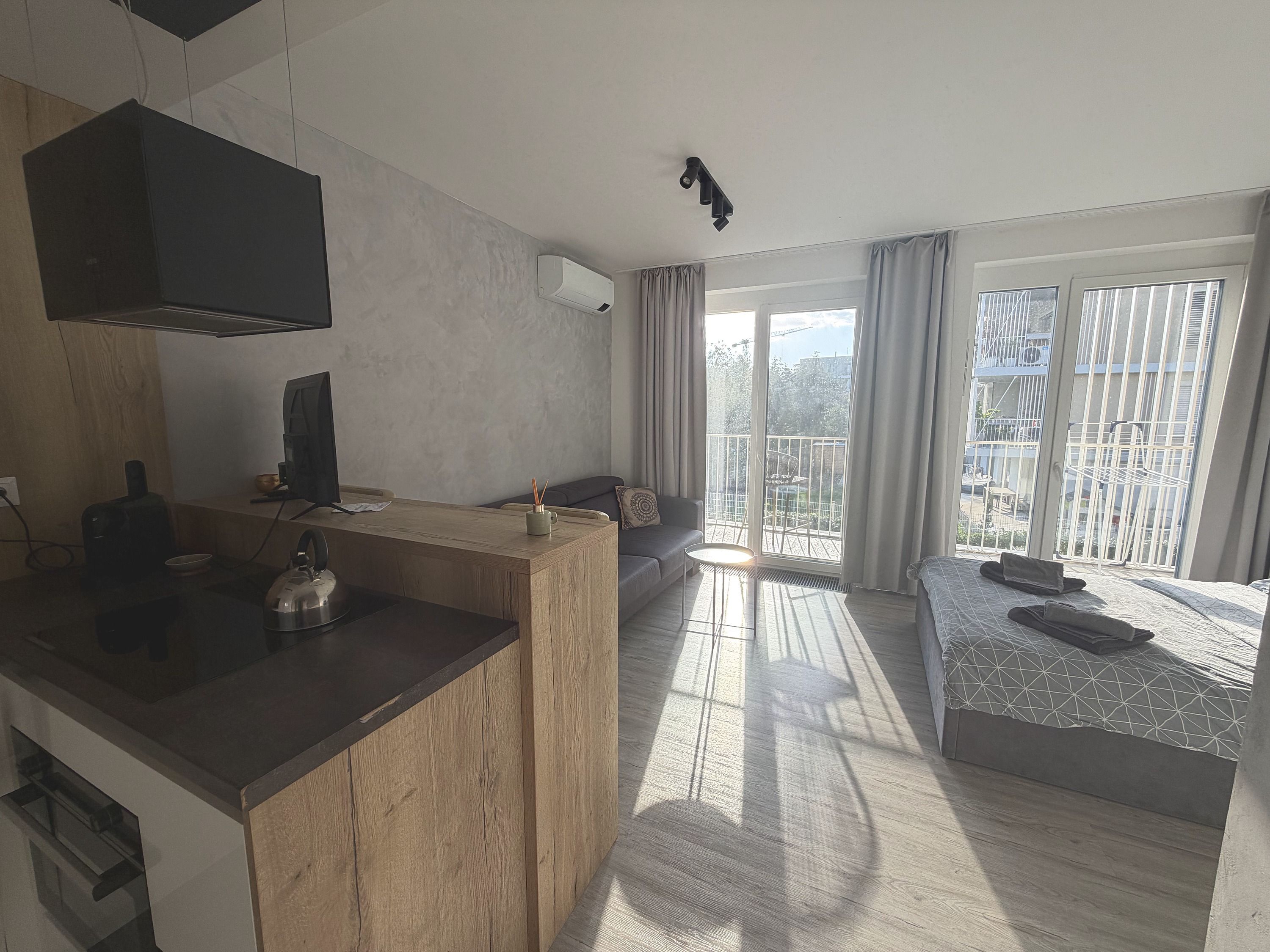 Une cuisine moderne avec un plan de travail en bois, une bouilloire et une machine à café. Le salon comprend un canapé et une petite table, et un balcon avec vue sur l’extérieur.