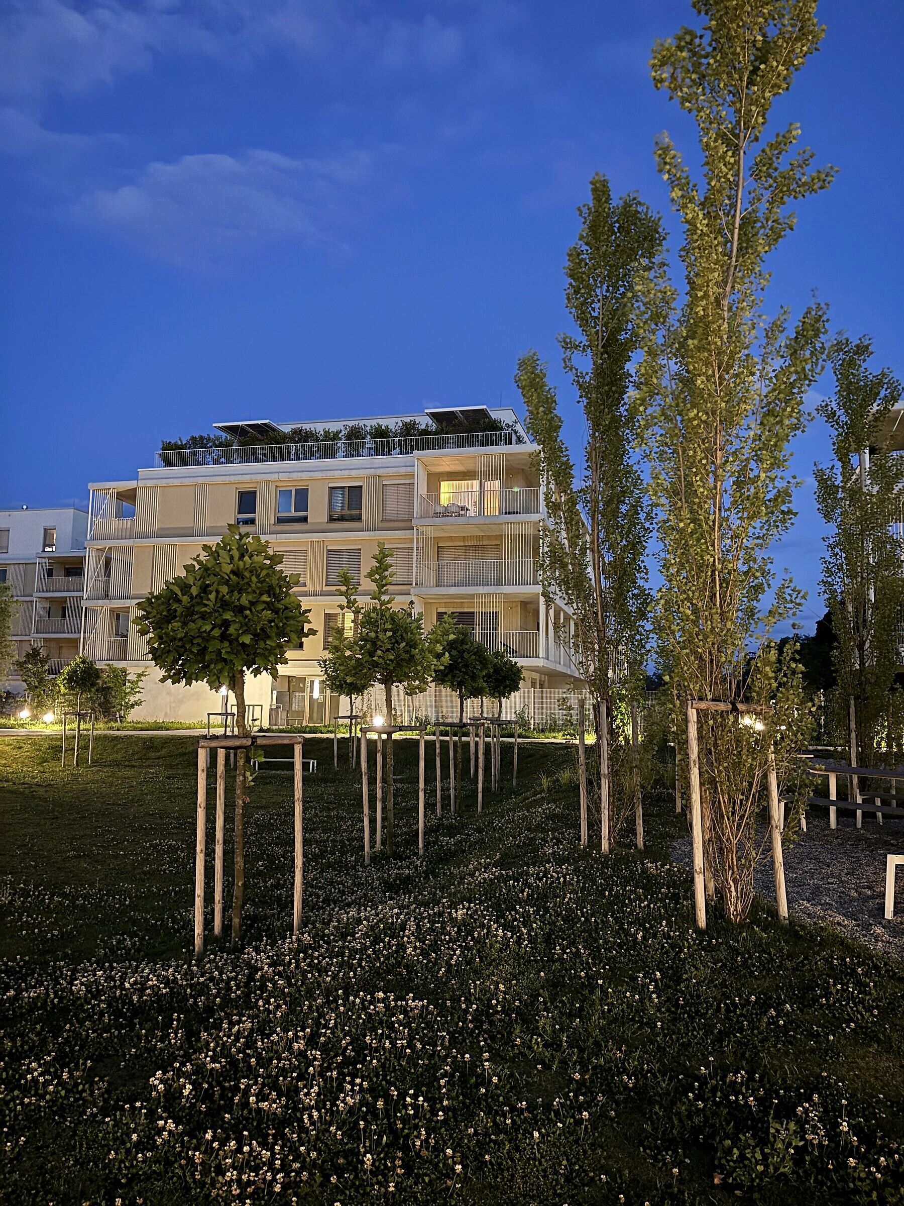 Un edificio residencial de varios pisos con balcones, rodeado por un jardín ajardinado con árboles y flores.