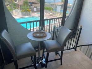 Ein Balkon mit Blick auf einen Pool, zwei Stühle, ein kleiner Tisch und ein Glas Wasser.