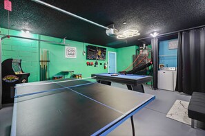 Une pièce équipée d’une table de ping-pong, d’une table de billard et d’une table de jeu de fléchettes. On y trouve également divers équipements sportifs et un téléviseur fixé au mur.