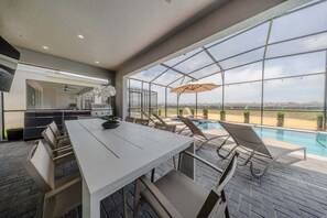 A modern outdoor kitchen area with a dining table and chairs, a poolside lounge area with sun loungers, and a large glass enclosure offering a view of the landscape. - Dream Retreat 8 BD/5 BA/Windsor Island - 3024 MD (Davenport)