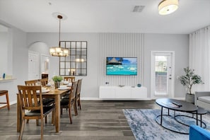 A modern living room with a dining area, a flat-screen TV mounted on the wall, a sofa, and a coffee table. - Near Epic! 3 BD/ 2 BA/ Vista Cay (4862-110 CA) (Orlando)