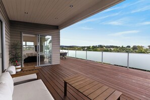 A modern balcony with wooden flooring, a sofa, and a glass door leading to an indoor space. - Sunset Vista - Mangawhai Heads Holiday Home (Mangawhai)
