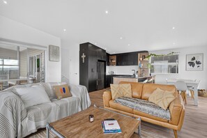 A modern living room with a sofa, a coffee table, and a kitchen area with dark cabinetry and a window. - Sunset Vista - Mangawhai Heads Holiday Home (Mangawhai)