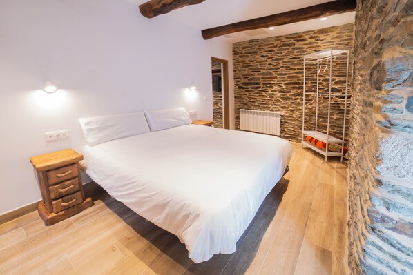 A bedroom with a large bed, wooden nightstands, a stone wall, and a hanging shoe rack. - TWO-BEDROOM PLUS APARTMENT (Castroverde)