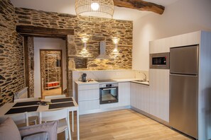 A modern kitchen with stone walls, a built-in oven, a refrigerator, and a dining area with a table and chairs. - TWO-BEDROOM PLUS APARTMENT (Castroverde)