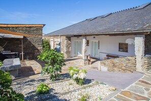 A stone building with a tiled roof, a small garden area with pebbles and plants, and a barbecue grill. - TWO-BEDROOM PLUS APARTMENT (Castroverde)