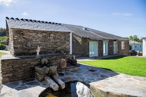A stone building with a sloped roof, a small water feature, and a grassy yard. - TWO-BEDROOM PLUS APARTMENT (Castroverde)