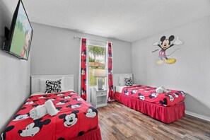 Ein Hotelzimmer mit zwei Betten, jeweils mit einem Mickey Mouse Bezug und einer Mickey Mouse Wanddekoration, sowie einem Fenster mit Vorhängen.