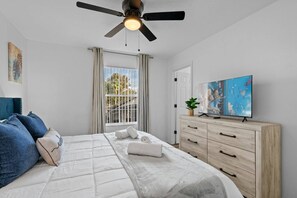 Ein Schlafzimmer mit Bett, Schminktisch, Deckenventilator, einem Fenster mit Vorhängen und einem Bild an der Wand.