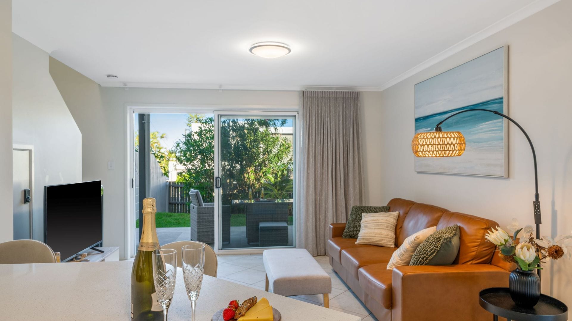 A modern living room with a sofa, a dining table with a bottle of champagne and glasses, and a large window with a view of greenery.
