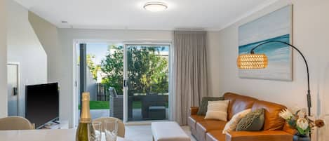 A modern living room with a sofa, a dining table with a bottle of champagne and glasses, and a large window with a view of greenery.