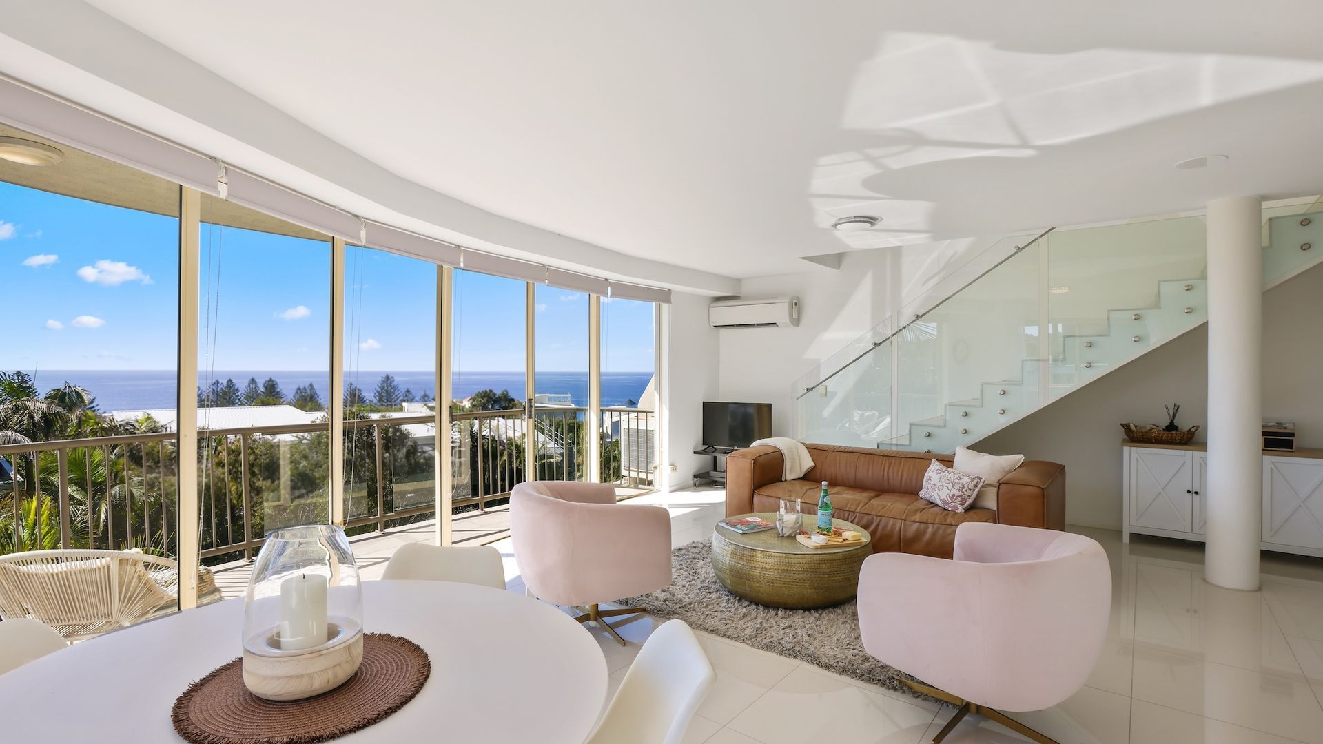 Ein modernes Wohnzimmer mit Panoramablick auf den Ozean, einer Glasstairnase und einem Essbereich mit gedecktem Tisch für zwei Personen.