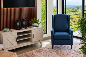 A modern living room with a blue armchair, wooden entertainment unit, and a large window with a view of greenery. - Alta Vista: Private 2BR Suite with Mountain & Sunset Views (Alajuela Province, San Ramon)