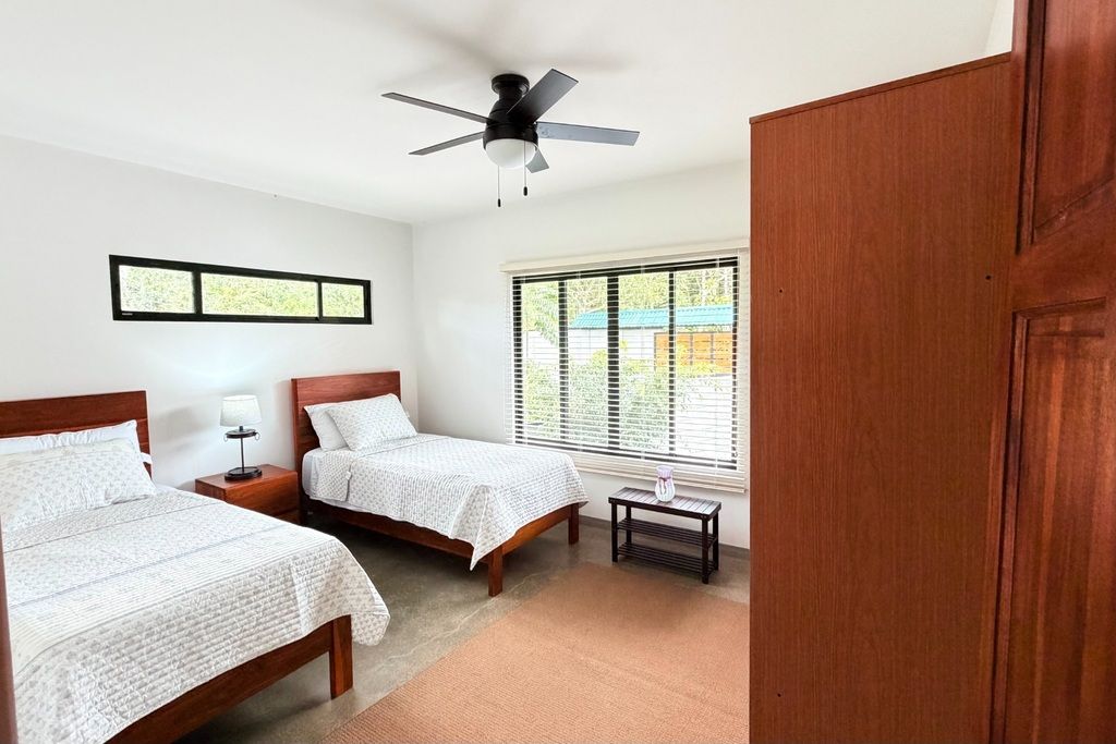 Une chambre avec deux lits, un ventilateur de plafond, une fenêtre avec des stores et une table de chevet avec une lampe.