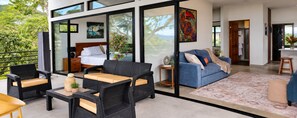 A modern interior with a bed, sofa, and dining area, featuring glass walls and a view of greenery. - Alta Vista: Private 2BR Suite with Mountain & Sunset Views (Alajuela Province, San Ramon)