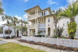A two-story house with a balcony, surrounded by a white picket fence, palm trees, and a clear sky. - 🌴Blue Skies & Tail Winds | Private Pool | 4 Bikes | Private Beach Access🌴 (Destin)