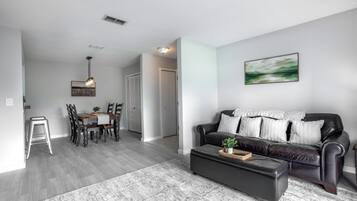 A modern living room with a black leather sofa, a dining table with chairs, and a hallway leading to a closet.
