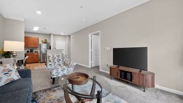 A modern living room with a flat-screen TV, a sofa, a coffee table, and a kitchen in the background.