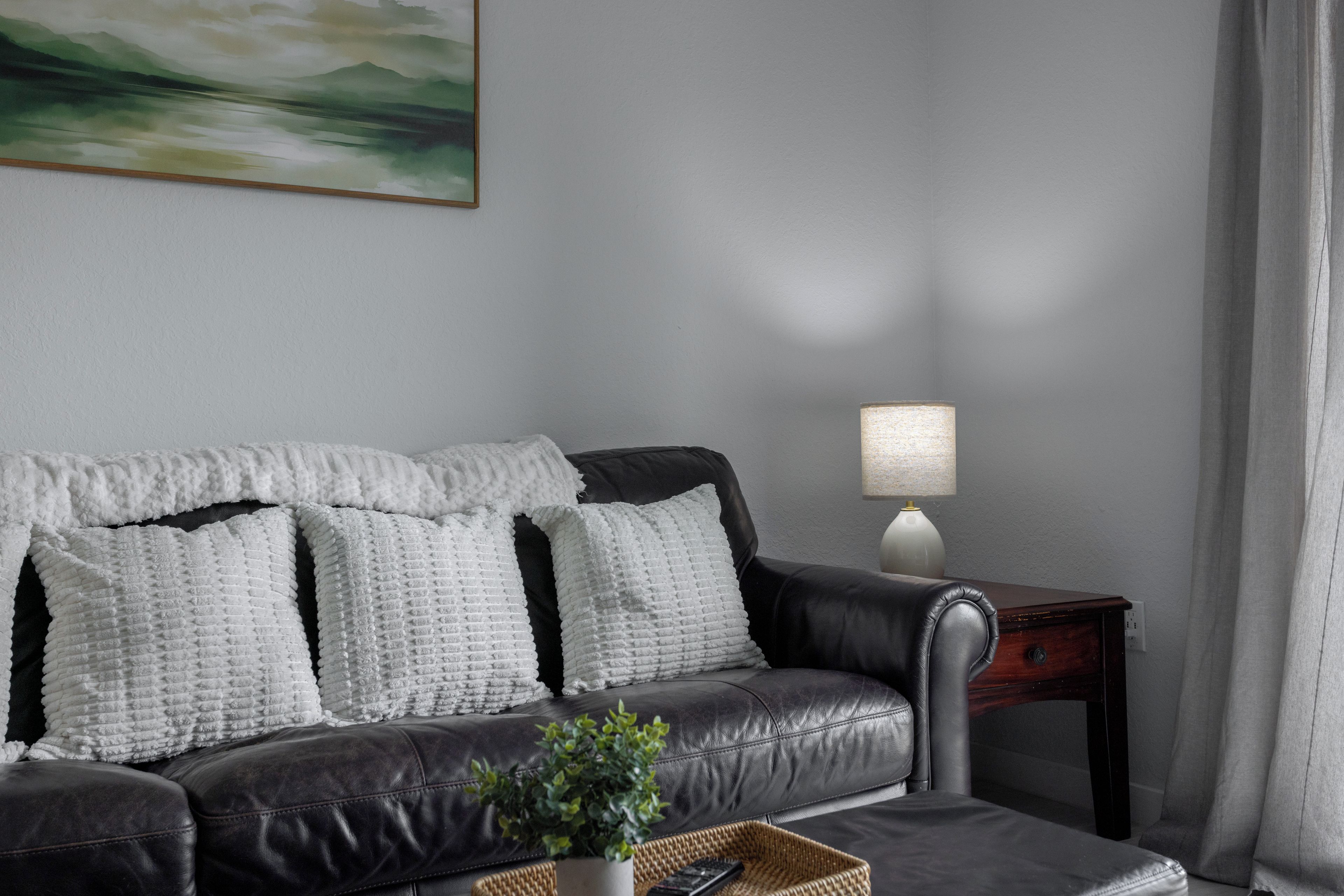 A modern living room with a leather sofa, white textured cushions, a side table with a lamp, and a painting on the wall.