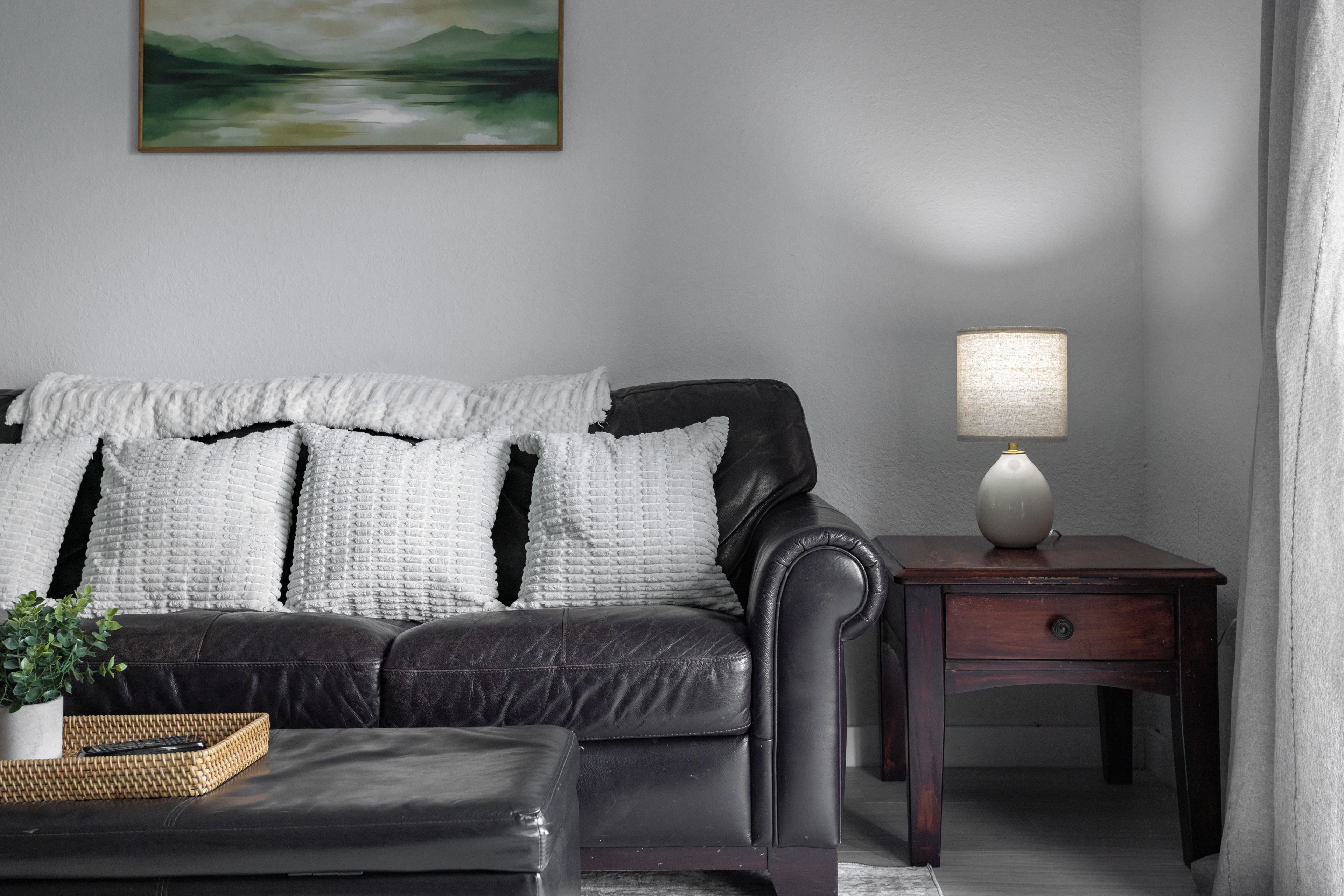 A dark leather sofa with white cushions, a small wooden side table with a lamp, and a framed landscape painting on the wall.