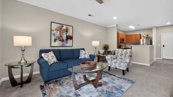 A living room with a sofa, two chairs, a coffee table, and a rug. There is a ceiling fan, a painting on the wall, and a kitchen area in the background.