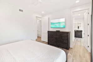 Ein Schlafzimmer mit einem großen Bett, einer dunkelholznen Kommode, einem flachbildfernseher an der Wand und einem durch eine offene Tür einsehbaren Badezimmer.