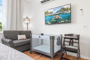 A room with a grey sofa, a baby playpen, a small table, and a wall-mounted TV displaying a sea turtle. - Updated | 1st Floor | Hilton Pool | Sunset Views (Waikoloa Village)