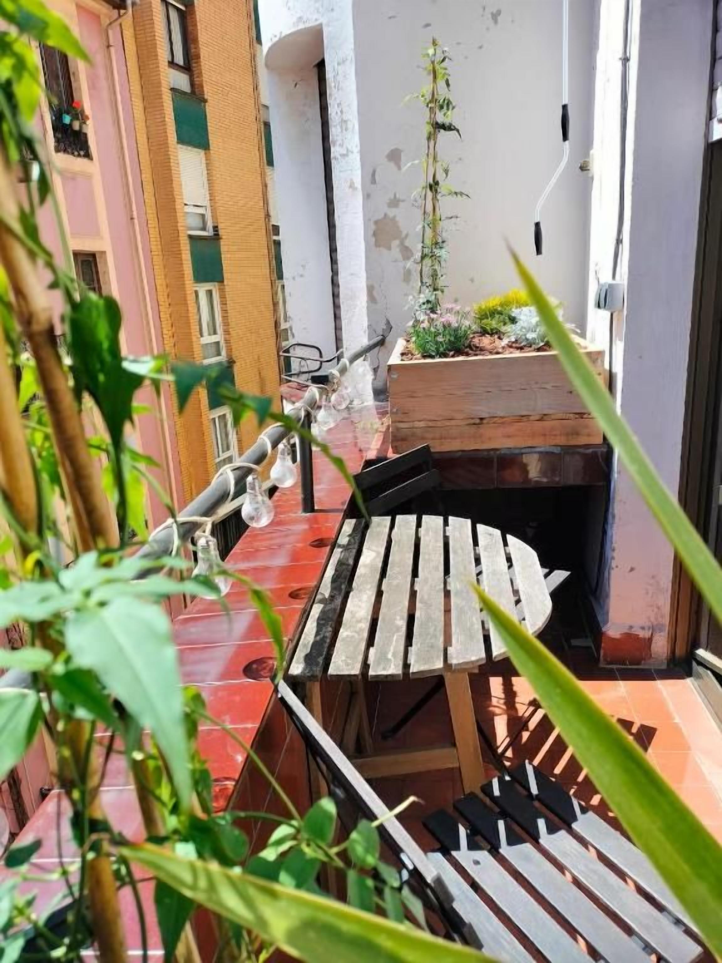 A narrow outdoor space with a wooden table and chairs, a small garden area, and a building with a white exterior and a brick wall.