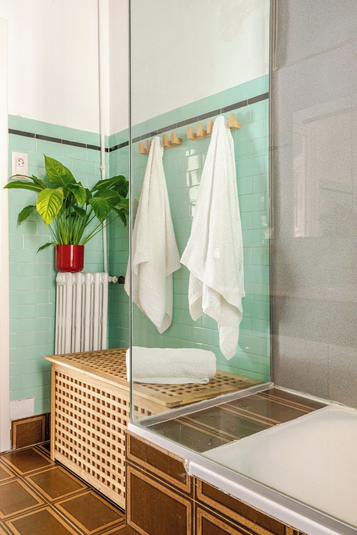A bathroom with a glass shower enclosure, a potted plant, a radiator, and a wooden laundry basket.